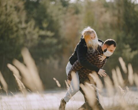 young man in mask carrying piggyback woman along spring forest